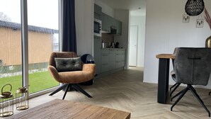 Een moderne woonkamer met een houten eettafel, een bruine fauteuil en een grijze stoel. In de achtergrond is een keuken zichtbaar met een magnetron en kastjes.