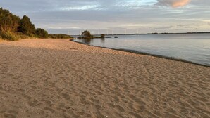 A sandy beach with a calm water body, trees, and wind turbines in the distance.