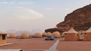 A desert landscape with multiple dome-shaped structures, a paved walkway, and a vehicle. - Bedouin life (Wadi Rum)