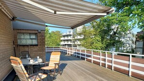 A covered outdoor seating area with a table and chairs, a brick wall, and a view of buildings and trees. - Die Insel Appartement 19 (Timmendorfer Strand)