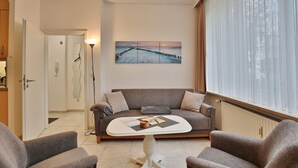 A modern living room with a grey sofa, a white coffee table, and a painting of a pier on the wall. - Die Insel Appartement 19 (Timmendorfer Strand)