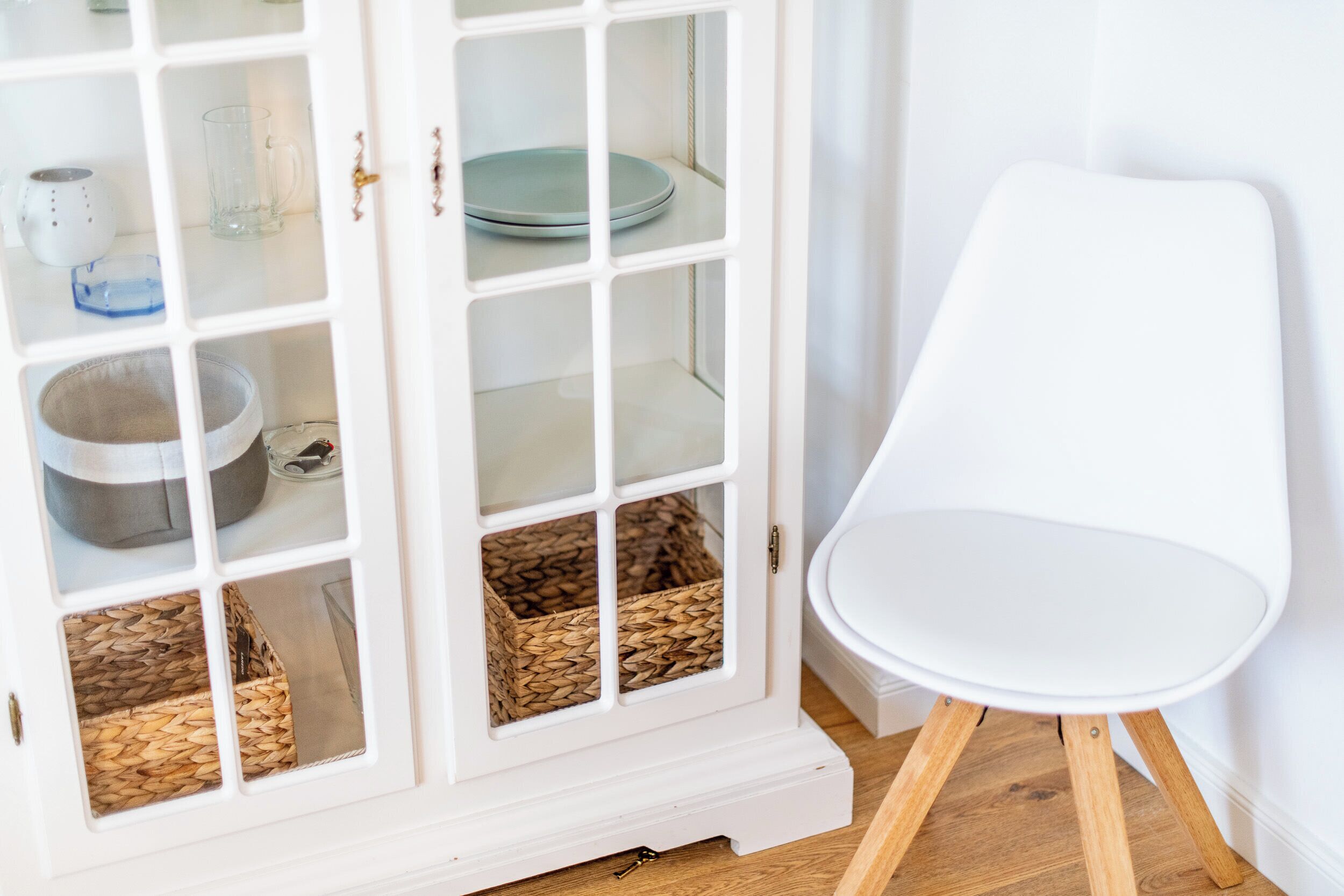 A white cabinet with glass doors containing baskets and a bowl, next to a white chair.