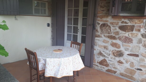 Un pequeño patio con una mesa redonda y dos sillas, un muro de piedra y una ventana con contraventanas.