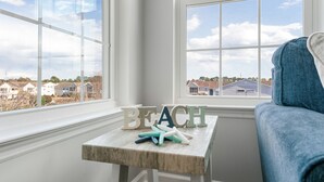 Une chambre d’angle avec vue sur les maisons, une petite table en bois sur laquelle sont posées des lettres formant le mot « PLAGE », et une chaise bleue.