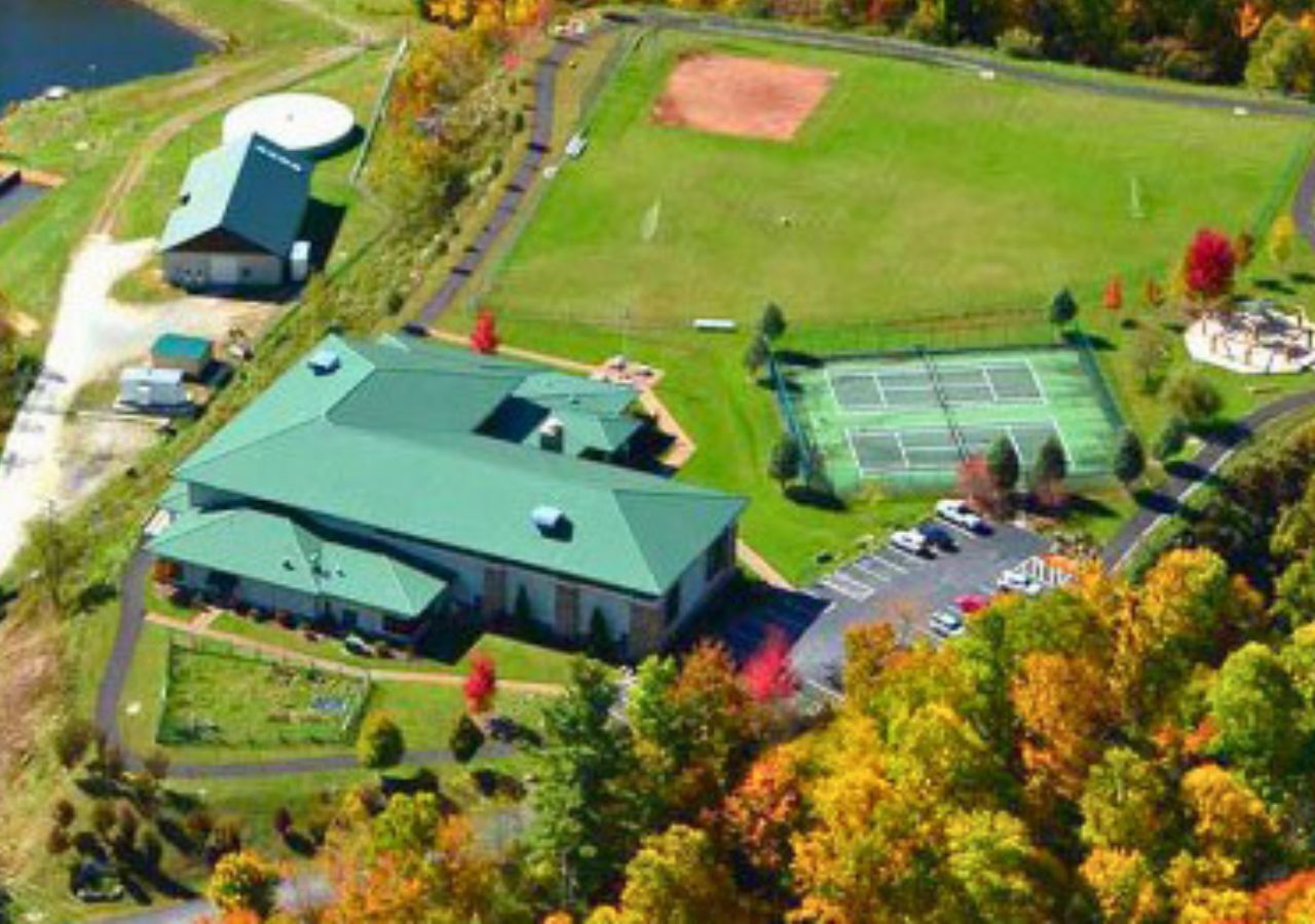 An aerial view of a large building with a green roof, surrounded by a tennis court, parking area, and a small lake.