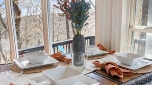Ett matbord dukat med vita, fyrkantiga tallrikar, skålar och en genomskinlig glasbringa. Bordet har en vävd bordstablett och en vas med torkade blommor.