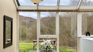 Une véranda avec une table et des chaises, un tableau encadré au mur et une vue sur un jardin et des collines.