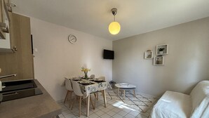 A compact living space with a dining area, a small round table, a sofa, a TV mounted on the wall, and a clock. - All-inclusive cottage between Moulins and Saint-Pourçain-sur-Sioule (Contigny)