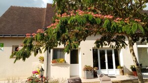 A building with a sloped roof, a covered patio with a chair, and a tree with pink flowers. - Charming Gîte with Private Garden, Close to Amboise, Perfect for Relaxation and Exploration (Pocé-sur-Cisse)