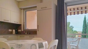 A kitchen with a dining table and chairs, a window with curtains, and a view of trees and buildings. - Air-Conditioned T2 with Parking, Bormes-les-Mimosas (Bormes-les-Mimosas)