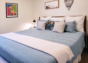 Ein ordentlich bezogenes Bett mit blauer und weißer Bettwäsche, ein kleiner Nachttisch mit Lampe und ein gerahmtes Bild von einem Hund an der Wand.