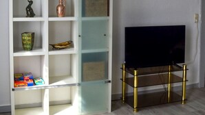 Ein Zimmer mit Holzboden, einem weißen Bücherregal mit verschiedenen Gegenständen, einem Fernsehgerät auf einem Schrank und einer Wanduhr.
