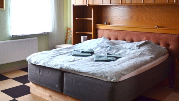 Ein Schlafzimmer mit Schachbrettmuster-Boden, einem Bett mit grauem Matratze, einem Holz-Kopfteil und einer hängenden Leuchte.