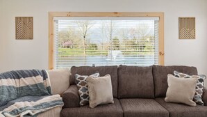 A living room with a brown sofa, a striped blanket, and a view of a garden through a window with blinds. - Pondview 4BR Cabin-Walk to Caverns, Hot Tub, Wi-Fi (Luray)