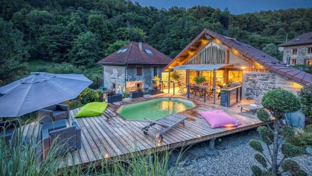 A wooden deck with a swimming pool, outdoor furniture, and a stone building with a sloped roof. - Le Chalet des Eaux Piscine (Revel)