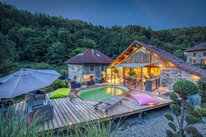 A wooden deck with a swimming pool, outdoor furniture, and a stone building with a sloped roof. - Le Chalet des Eaux Piscine (Revel)