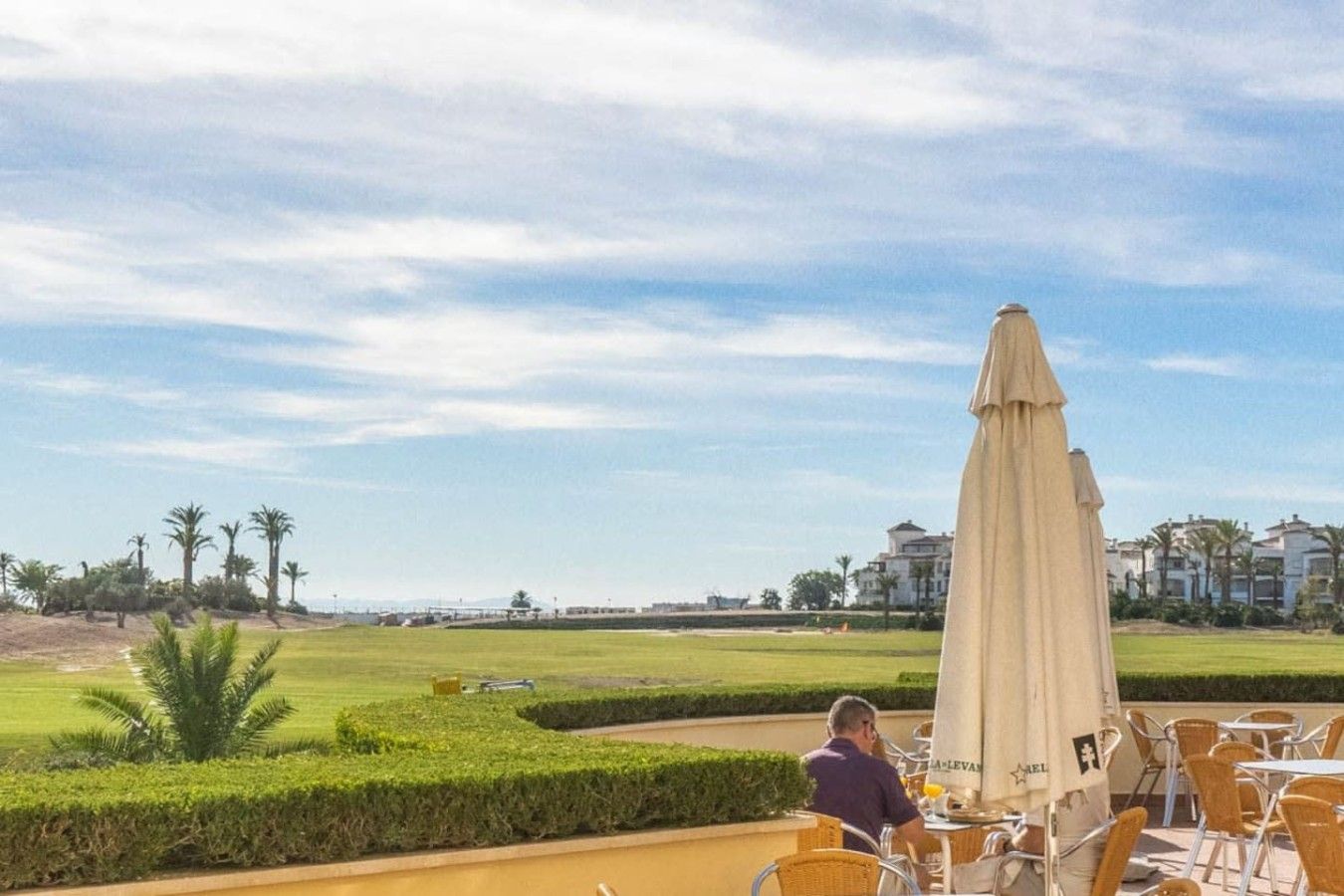 Un homme est assis à une table en plein air, avec vue sur un terrain de golf et des bâtiments en arrière-plan.
