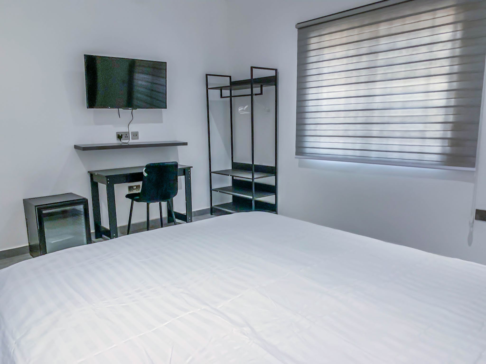 Habitación de hotel con una cama grande, un escritorio con silla, un televisor montado en la pared y una ventana con persianas.