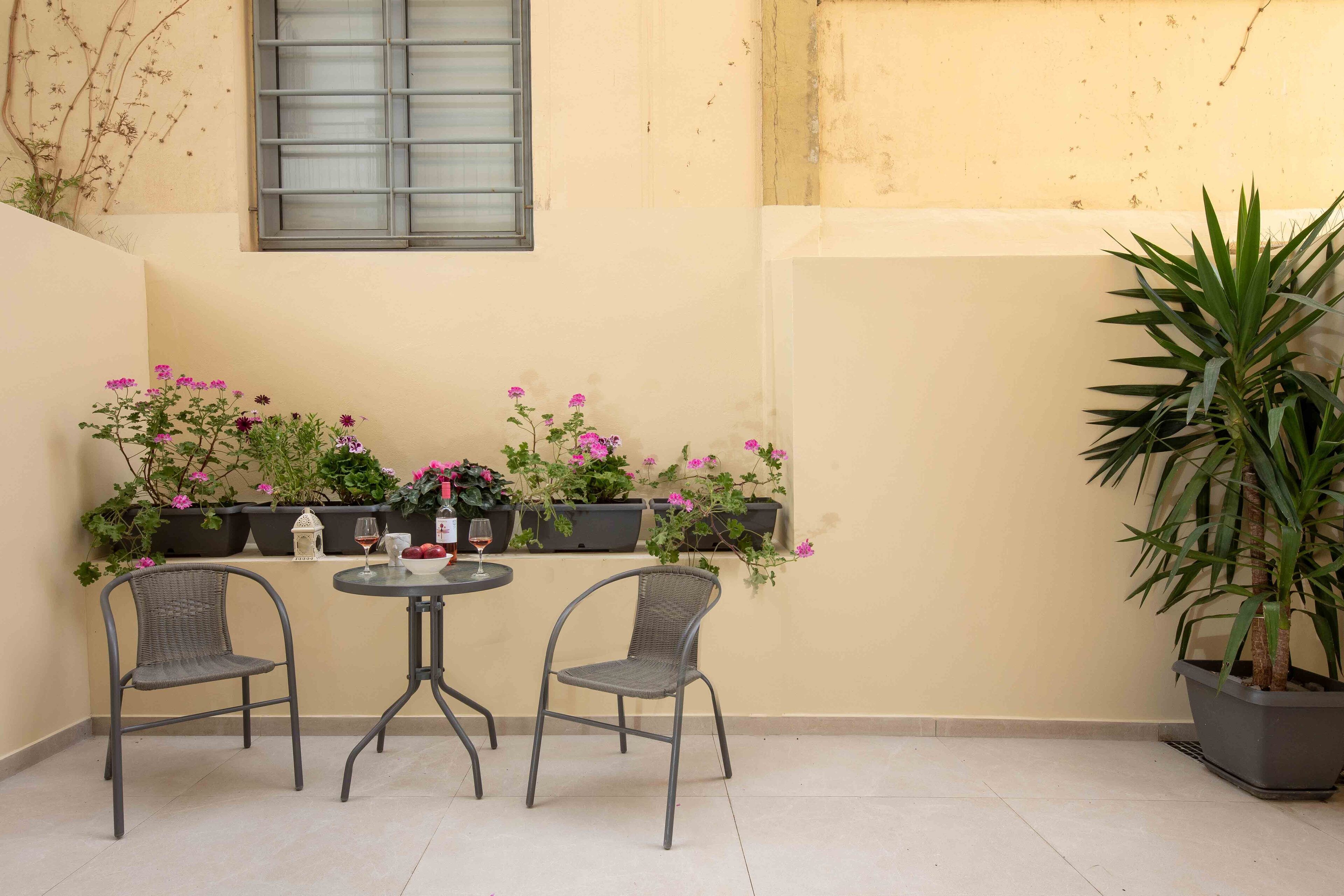 A small patio area with two wicker chairs and a round table, surrounded by potted plants and flowers.