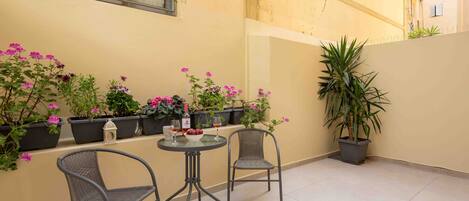 Un pequeño patio con dos sillones de mimbre, una mesa redonda y macetas con flores rosadas.