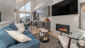 A modern living room with a blue sofa, a fireplace, a flat-screen TV, and a kitchen in the background. - Skyview Retreat - Pool, Hot Tub &  Parking (Canmore)