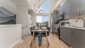 A modern kitchen with grey cabinets, a dining table set for four, and a view of mountains through large windows. - Skyview Retreat - Pool, Hot Tub &  Parking (Canmore)
