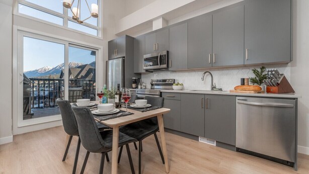 A modern kitchen with grey cabinets, stainless steel appliances, and a dining area with a table and chairs. - Skyview Retreat - Pool, Hot Tub &  Parking (Canmore)