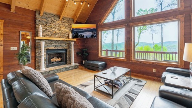 A cozy living room with a stone fireplace, leather sofas, and large windows offering a view of the outdoors. - A Mtn Frame of Mind- Mountain View, Hot Tub, Game Room, Pet Friendly (Seven Devil's)