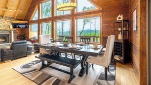 A spacious dining area with a wooden table and chairs, large windows offering a view of the outdoors, and a stone fireplace. - A Mtn Frame of Mind- Mountain View, Hot Tub, Game Room, Pet Friendly (Seven Devil's)
