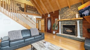 A spacious living room with a black leather sofa, wooden staircase, stone fireplace, and a view of the outdoors. - A Mtn Frame of Mind- Mountain View, Hot Tub, Game Room, Pet Friendly (Seven Devil's)