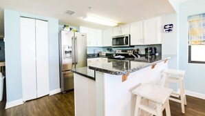 Cocina compacta con armarios blancos, encimera de granito, electrodomésticos de acero inoxidable y una pequeña isla con taburetes.