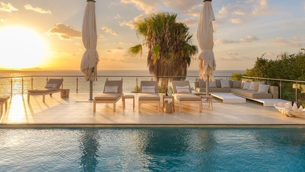Área da piscina com espreguiçadeiras e guarda-sóis, com vista para o mar ao pôr do sol.