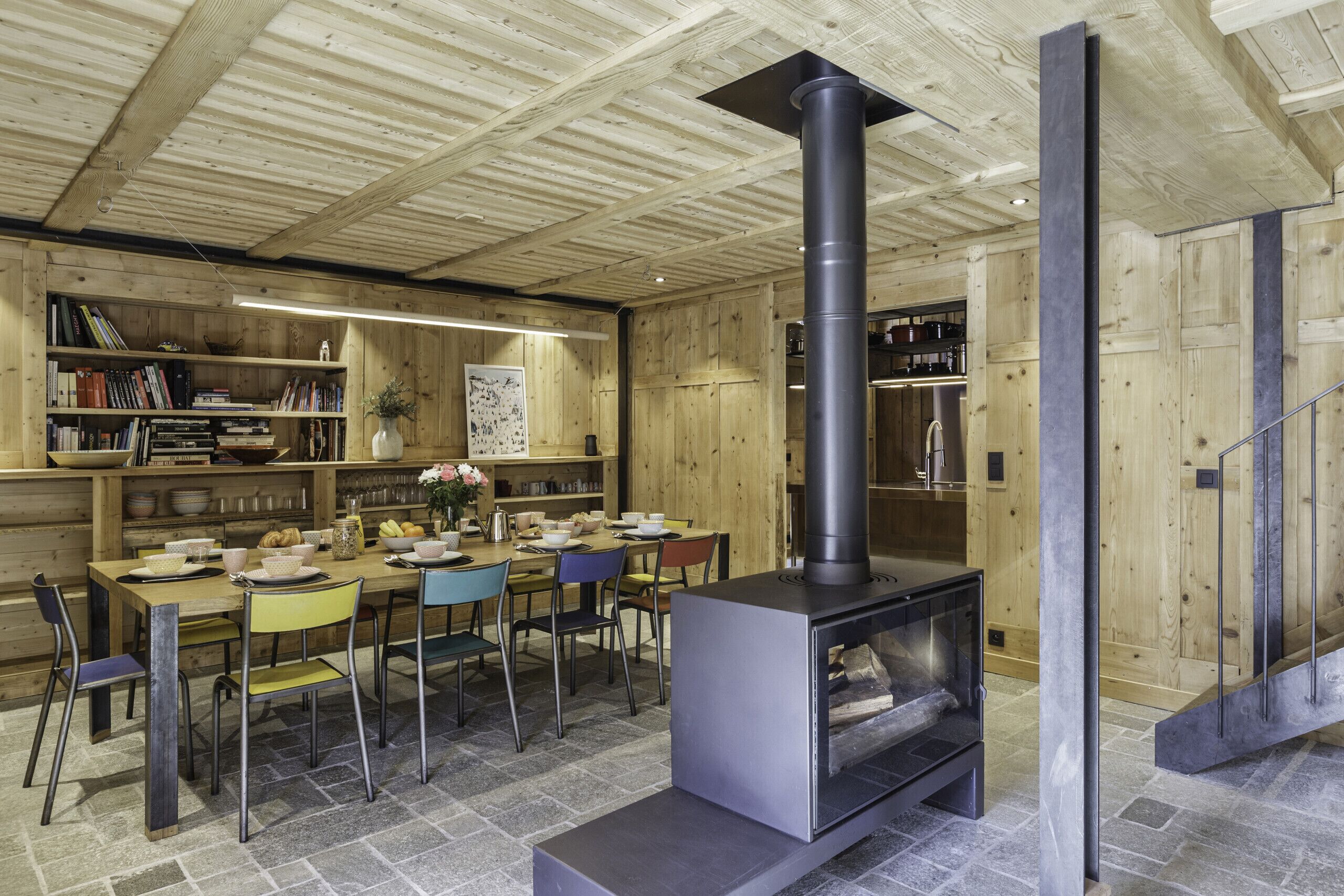 Een gezellig houten interieur met een centraal gelegen open haard, een eethoek met stoelen en een tafel, en planken met boeken en decoratieve objecten.