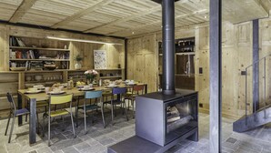 A cozy wooden interior with a central fireplace, a dining area with chairs and a table, and shelves with books and decorative items.