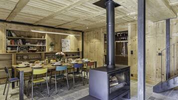 Een gezellig houten interieur met een centraal gelegen open haard, een eethoek met stoelen en een tafel, en planken met boeken en decoratieve objecten.