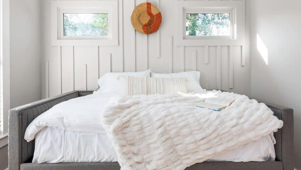 A bed with white bedding and a white throw blanket, a gray headboard, and two windows with a view of greenery. - Zen Den (Atlanta)