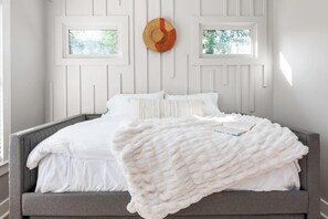 A bed with white bedding and a white throw blanket, a gray headboard, and two windows with a view of greenery. - Zen Den (Atlanta)