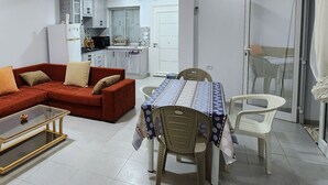Una sala de estar con sofá, mesa de centro y mesa de comedor con sillas. Se ve una cocina al fondo, con armarios y un refrigerador.
