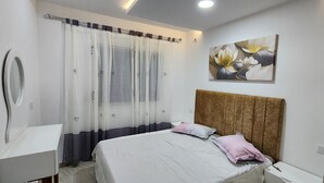 Un dormitorio con una cama, una mesita de noche, un espejo, un ventanal con cortinas translúcidas y una obra de arte colgada en la pared.
