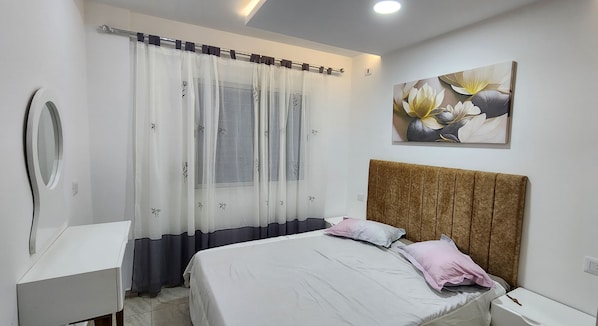 Un dormitorio con una cama, una mesita de noche, un espejo, un ventanal con cortinas translúcidas y una obra de arte colgada en la pared.