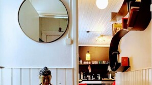 Une statue se trouve dans un couloir, avec un miroir au-dessus, qui mène à un espace cuisine doté de chaises rouges et d’un tapis à motifs.