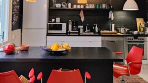 Une cuisine moderne avec des meubles noirs, un îlot central sur lequel se trouve un panier de fruits, des chaises rouges et des luminaires suspendus.