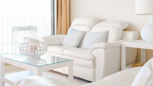 Un sofá de cuero blanco con dos almohadas, una mesa de centro de vidrio y una lámpara blanca sobre una mesita auxiliar blanca.