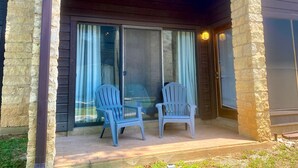 A covered patio area with two chairs and a table, stone walls, and a sliding glass door with curtains. - River City Resorts: B115 : Free Tubes with Rental! (New Braunfels)