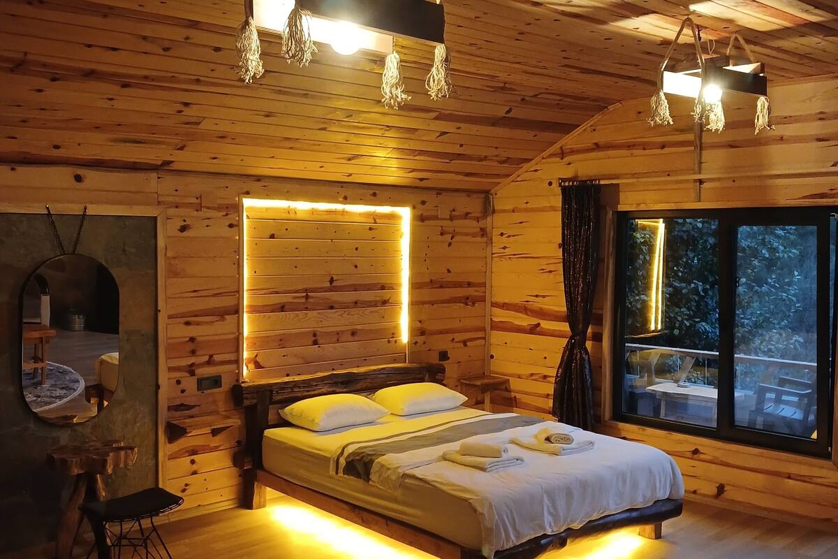 Camera da letto di un accogliente chalet in legno, con un letto grande, lampade sui comodini e vista dalla finestra.