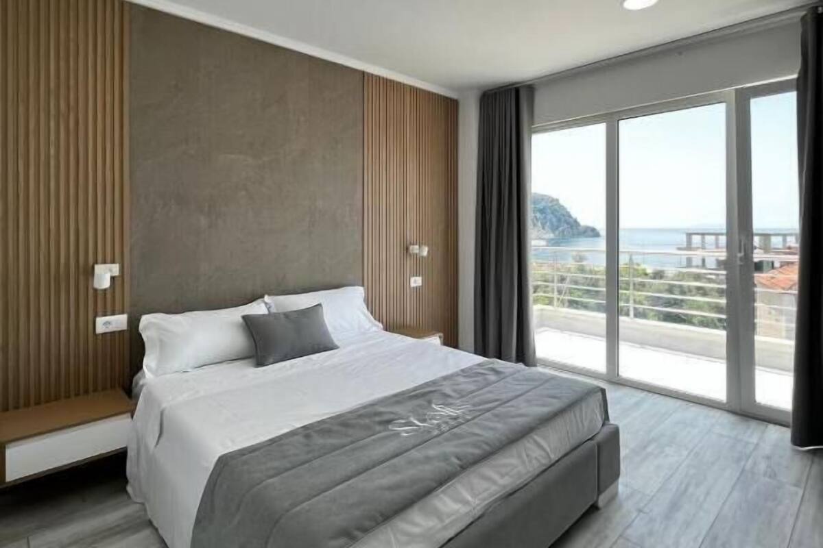 A modern bedroom with a large bed, a wooden panel wall, a large window with a view, and a ceiling light.