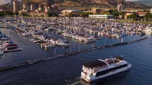 A marina filled with numerous boats docked in calm waters, with a large building and hills in the background. - The Central | Downtown + Rooftop Patio (Kelowna)