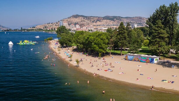 A beach with clear waters, a sandy shore, and a backdrop of buildings and hills. - The Central | Downtown + Rooftop Patio (Kelowna)