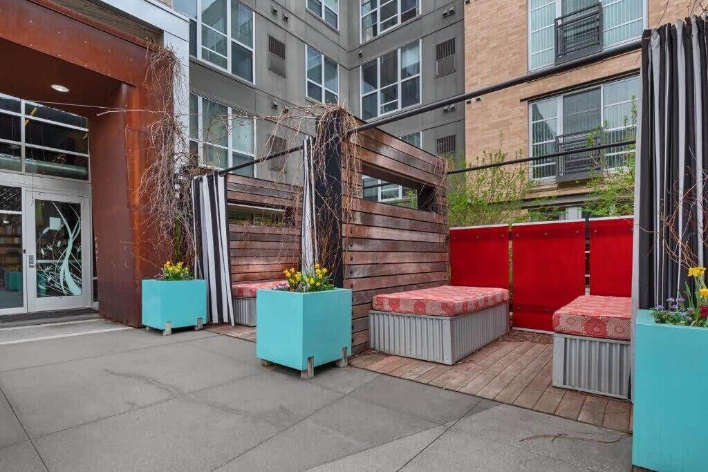 An outdoor patio area with wooden benches and planters, a red fence, and a building with large windows in the background.