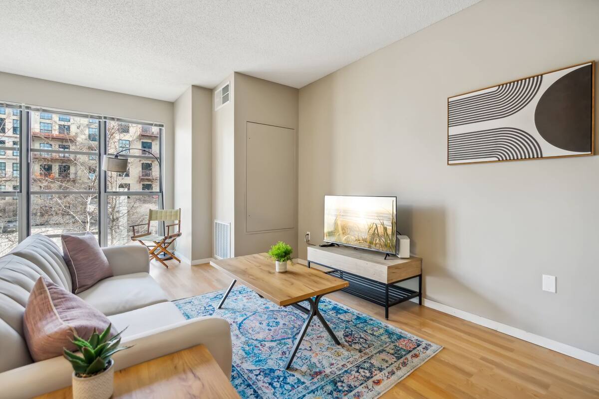 Un salon moderne avec un canapé, une table basse, un téléviseur et de grandes fenêtres donnant sur des immeubles.